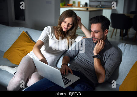 Young couple sitting on sofa in evening looking at laptop Stock Photo