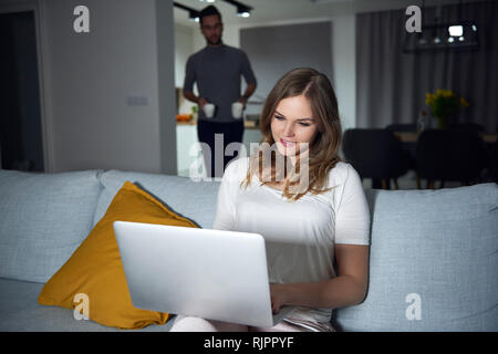Young man bringing coffee to girlfriend sitting on sofa using laptop in evening Stock Photo