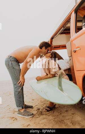 Young surfing couple kissing by recreational vehicle on beach,  Jalama, Ventura, California, USA Stock Photo