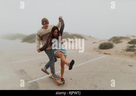 Young couple holding hands sharing a skateboard in beach carpark, Jalama, California, USA Stock Photo