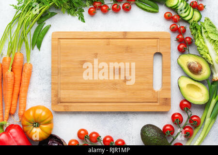 Fresh raw ingredients arranged in a circle with chopping board in the middle tomatoes carrots lettuce pepper avocado on the white table, top view, selective focus Stock Photo