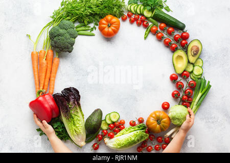 Childs hands holding composition from tomatoes cucumbers lettuce pepper avocado carrots broccoli peas in a circle frame on the white table, top view Stock Photo