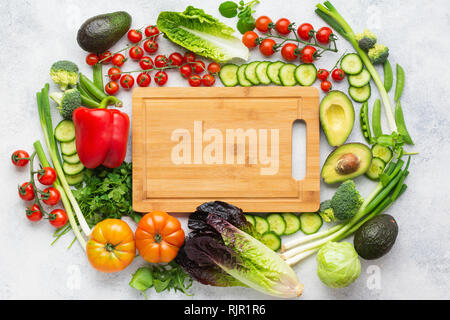 Fresh ingredients arranged in a circle with chopping board in the middle, tomatoes cucumbers lettuce pepper avocado parsley spring peas on white table Stock Photo