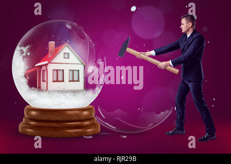 A businessman with a hammer in his hands crashing a crystal ball with a little house inside on a purple background with a few flecks of snow. Building Stock Photo