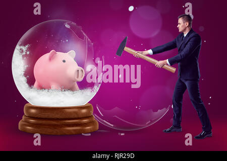 A businessman with a hammer in his hands crashing a big crystal ball with a piggy bank inside on a purple background with a few flecks of snow. Financ Stock Photo