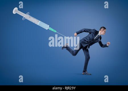 A side view of a businessman running from a huge syringe floating in the air on a blue background. Fear of vaccines. Refusal of vaccines and vaccine d Stock Photo