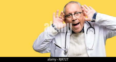 Handsome senior doctor man wearing medical coat Trying to hear both hands on ear gesture, curious for gossip. Hearing problem, deaf Stock Photo