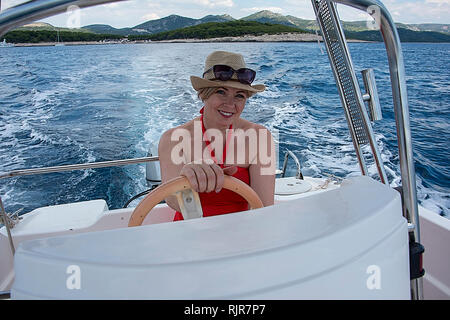 Croatia, Hvar - June 2018: Women in mid 40's driving a small boat.  She wears a red dress and hat hat and sunglasses on her head. Coastline is visible Stock Photo