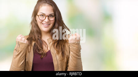 Beautiful plus size young woman wearing a dress and leather jacket over isolated background celebrating surprised and amazed for success with arms rai Stock Photo