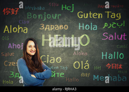 Cheerful woman student holding arms crossed in front of a huge blackboard written with the word hello translated in different languages and colors. Op Stock Photo