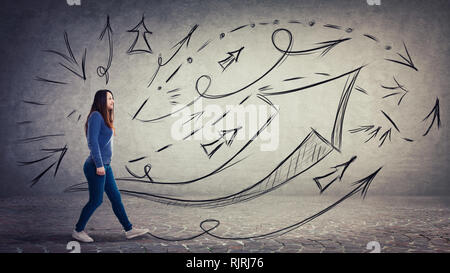 Side view full length portrait, positive young woman walking over grey wall background with different arrows and lines leading ahead. Choosing the cor Stock Photo