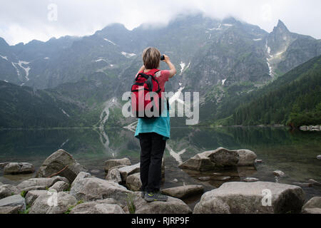 A woman stands on the rocky bank of Morskie Oko lake (Eye of the Sea) taking a photo with her cell phone in the Tatra National Park,Lesser Poland Vovo Stock Photo