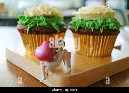 Closed up mini angel holding pink big heart figurine with a pair of flower shaped whipped cream chocolate cupcakes in background Stock Photo