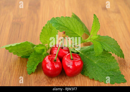 Three cherries with stems and lemon balm leaves on dark wooden surface Stock Photo