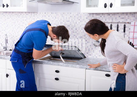 Woman Looking At Serviceman In Uniform Fixing Induction Stove In The Kitchen Stock Photo