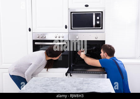 Smiling Young Woman Looking At Technician Fixing Oven In Kitchen Stock Photo