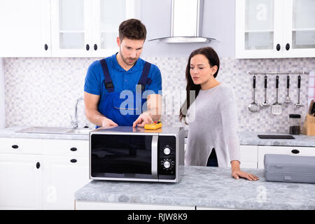 Pretty Woman Standing Near Repairman Repairing Oven Using Digital Multimeter In Kitchen Stock Photo