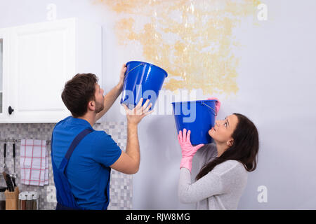 Young Woman And Repairman With Blue Bucket Collecting Water From Damaged Ceiling In Kitchen Stock Photo