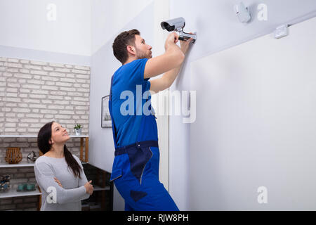 Smiling Woman Looking At Male Technician Adjusting CCTV Camera On Wall With Screwdriver Stock Photo