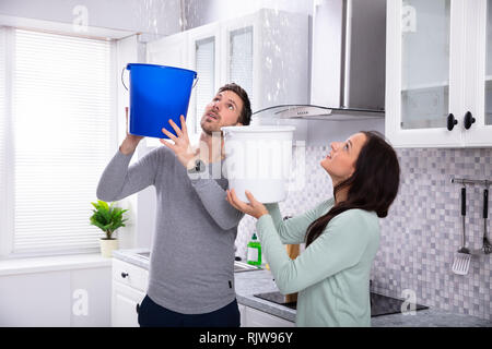 Close-up Of A Worried Young Couple Collecting Water Leaking From Ceiling In The Blue Bucket Stock Photo