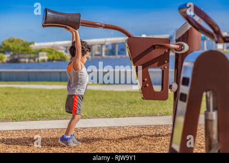 The little boy stretches high to reach the adult shoulder pads at the waterfront outdoor gym. Stock Photo