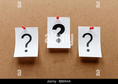 Three question marks pinned to a wooden notice board with a bold one in the centre printed on white paper cards Stock Photo