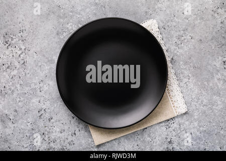 Empty ceramic plate in black with a napkin on gray concrete table, top view Stock Photo