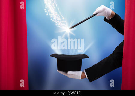 Magician Doing Magic Trick With Illuminated Wand And Hat Behind Curtain Stock Photo