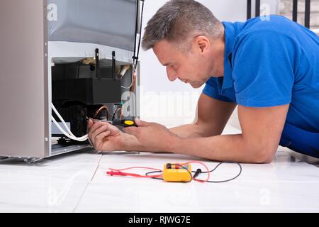 Male Serviceman In Overall Working On Fridge With Screwdriver In Kitchen Stock Photo