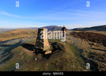 Walker at the  OS Trig Point at the summit Cairn of Ling Fell, Wythop Moss, Lake District National Park, Cumbria, England, UK Stock Photo