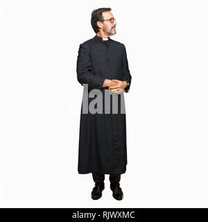 Middle age priest man wearing catholic robe smiling looking to the side with arms crossed convinced and confident Stock Photo