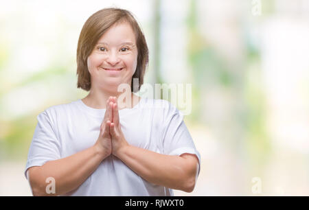 Young adult woman with down syndrome over isolated background praying with hands together asking for forgiveness smiling confident. Stock Photo