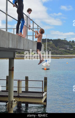 Boy hanging from metal railing of concrete jetty ready to jump into calm sea. Stock Photo