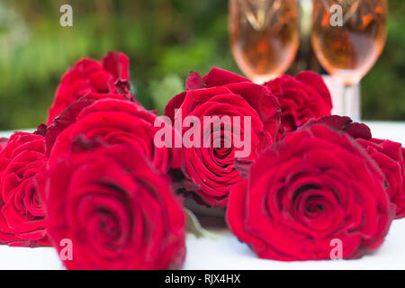 roses red lying scattered on a white table outdoors with two glasses filled with champagne or sparkling wine in the background ready for celebration Stock Photo