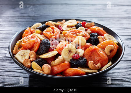 close-up of dried organic Fruits and Nut Mix - banana slices, apricots, raisins, prunes, cherries and cashew on a black plate on a wooden table, healt Stock Photo