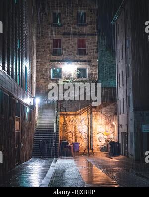 A singular figure about to climb some stairs down an alleyway in the city centre of Edinburgh, Scotland during the dark evening. The wet weather pours Stock Photo
