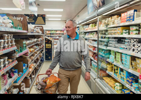 Senior man is walking down an aisle at a supermarket with a basket of shopping. Stock Photo