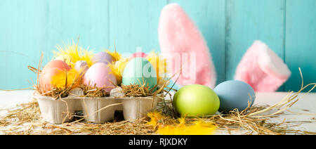 Pastel colored easter eggs in a egg box with hay and feathers as a nest and rabbit ears behind against a wooden turquoise background. Banner size Stock Photo