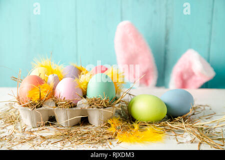 Pastel colored easter eggs in a egg box with hay and feathers as a nest and rabbit ears behind against a wooden turquoise background Stock Photo