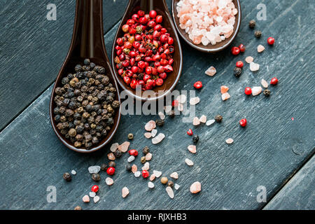 Top view of three spoons with rough pink salt, black and pink peppercorns on dark wooden background Stock Photo