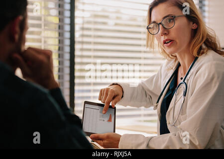 Confident female doctor talking with patient with test results on tablet. Doctor and patient talking about the treatment in hospital. Stock Photo