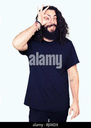 Young man with long hair and beard wearing heavy metal black outfit doing ok gesture with hand smiling, eye looking through fingers with happy face. Stock Photo