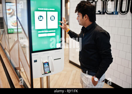 Indian man customer at store place orders and pay through self pay floor kiosk for fast food, payment terminal. Make a choise of language on screen. Stock Photo