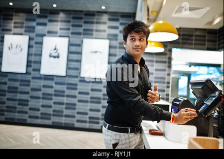 Indian man holding pay by credit card at cash desk with order screen and payment terminal in food cafe. Stock Photo