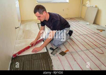 Young worker tiler installing ceramic tiles using lever on cement floor with heating red electrical cable wire system. Home improvement, renovation an Stock Photo