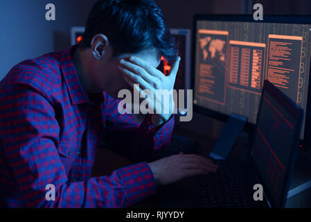 An anonymous male hacker covers his face with hand while committing a crime to hack the system on the background of screens in the dark. The concept o Stock Photo