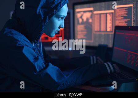 Girl hacker in a hood typing program code while committing a cybercrime hacking a system firewall on the background of screens in the dark under neon  Stock Photo