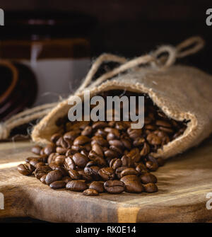 Closeup of coffee beans spilled from a burlap bag onto a wooden board with a dark rustic background Stock Photo