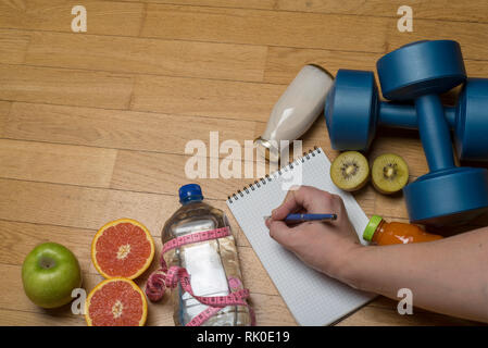 Training, exercise, cheerfulness and health - two plastic dumbbells, mineral water with juice, fruit and hand writes in a notebook on the wooden floor Stock Photo