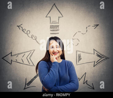 Ingenious young woman thinking to choose a direction as arrows on the gray wall are pointed to different directions ways. Difficult choice, making rig Stock Photo